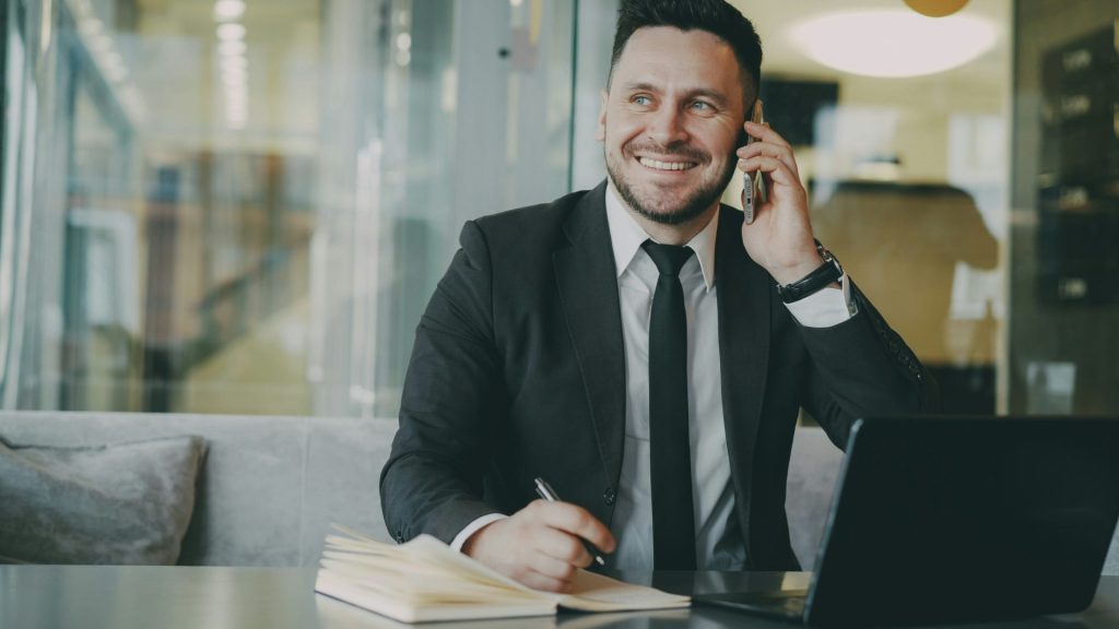 Un homme en costume parle au téléphone à son bureau avec un ordinateur portable.